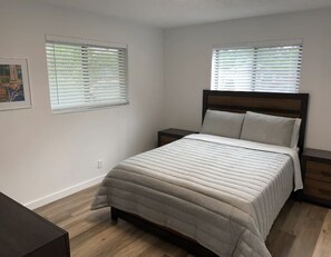 Une chambre à coucher avec un lit, deux tables de chevet et deux fenêtres équipées de stores.