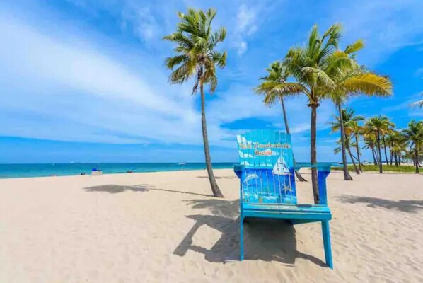 Une chaise de plage avec une pancarte indiquant «
Plage de Fort Lauderdale
» sur une plage de sable, bordée de palmiers et surplombée d’un ciel bleu clair.
