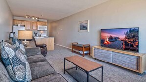 A living room with a sofa, a coffee table, and a TV. There is a kitchen in the background.