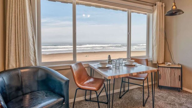 A room with a large window overlooking the beach, a wooden table with chairs, a leather chair, and a small cabinet.