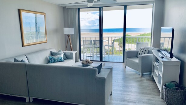 Een moderne woonkamer met een grijze bank, een salontafel, een tv-meubel en een balkon met uitzicht op het strand.