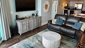 Een woonkamer met een grote flatscreen televisie, een leren bank met decoratieve kussens, een ronde salontafel en een kamerkleed met een patroon.