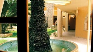 A stone fountain with water flowing into a circular pool, surrounded by a stone wall and a garden area. - Hotel Arrecife de Coral (San Cristóbal de las Casas)