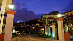 A well-lit entrance with a pathway leading to a building with multiple floors and a visible flag. - Hotel Arrecife de Coral (San Cristóbal de las Casas)