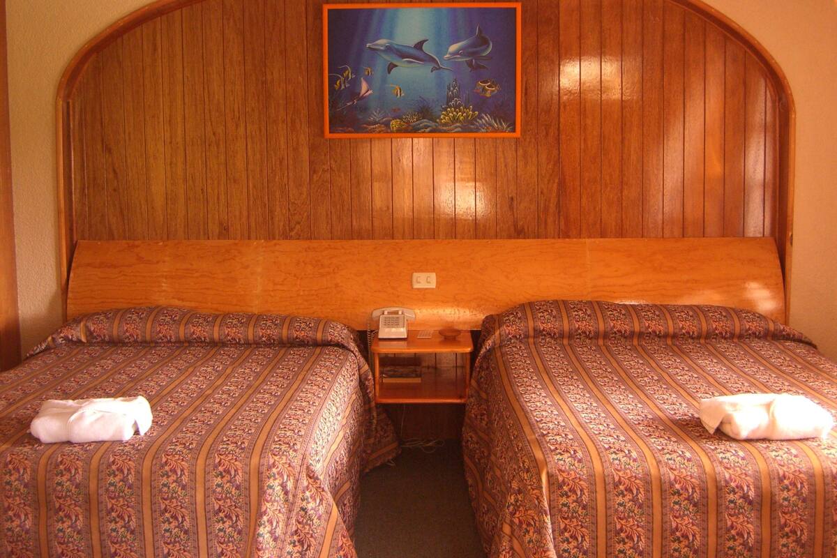 A hotel room with a wooden headboard, two beds with patterned bedding, a small table with a telephone, and an underwater painting.