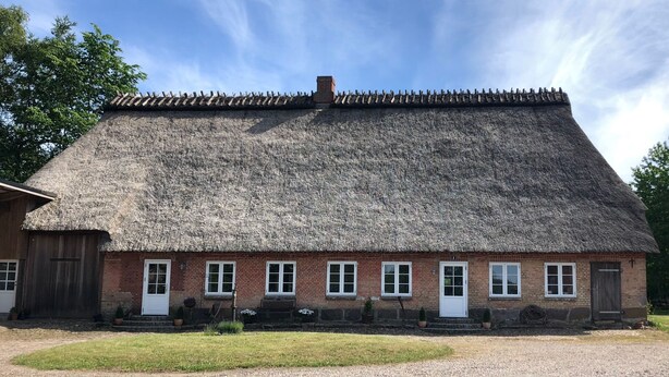 Ein traditionelles Gebäude mit Strohdach, Backsteinfassade, mehreren Fenstern und einem Schotterweg.