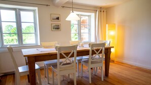 Ein Essbereich mit Holztisch und weißen Stühlen, großen Fenstern mit Vorhängen und einer Heizung.