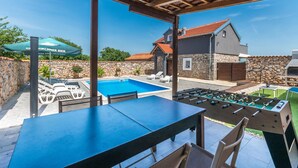 Ein Poolbereich mit Tischfußball, Liegestühlen und Blick auf ein Haus mit einer Steinmauer und einem roten Ziegeldach.