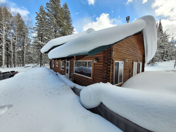 A log cabin with a snow-covered roof and windows, surrounded by a snowy landscape and trees.