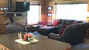 A log cabin interior with a wooden dining area, a sofa, and a television mounted on the wall. - Mighty Moose Log Cabin, 20 Minutes to W Yellowstone, Fishing, Sleeps 10. (Island Park)