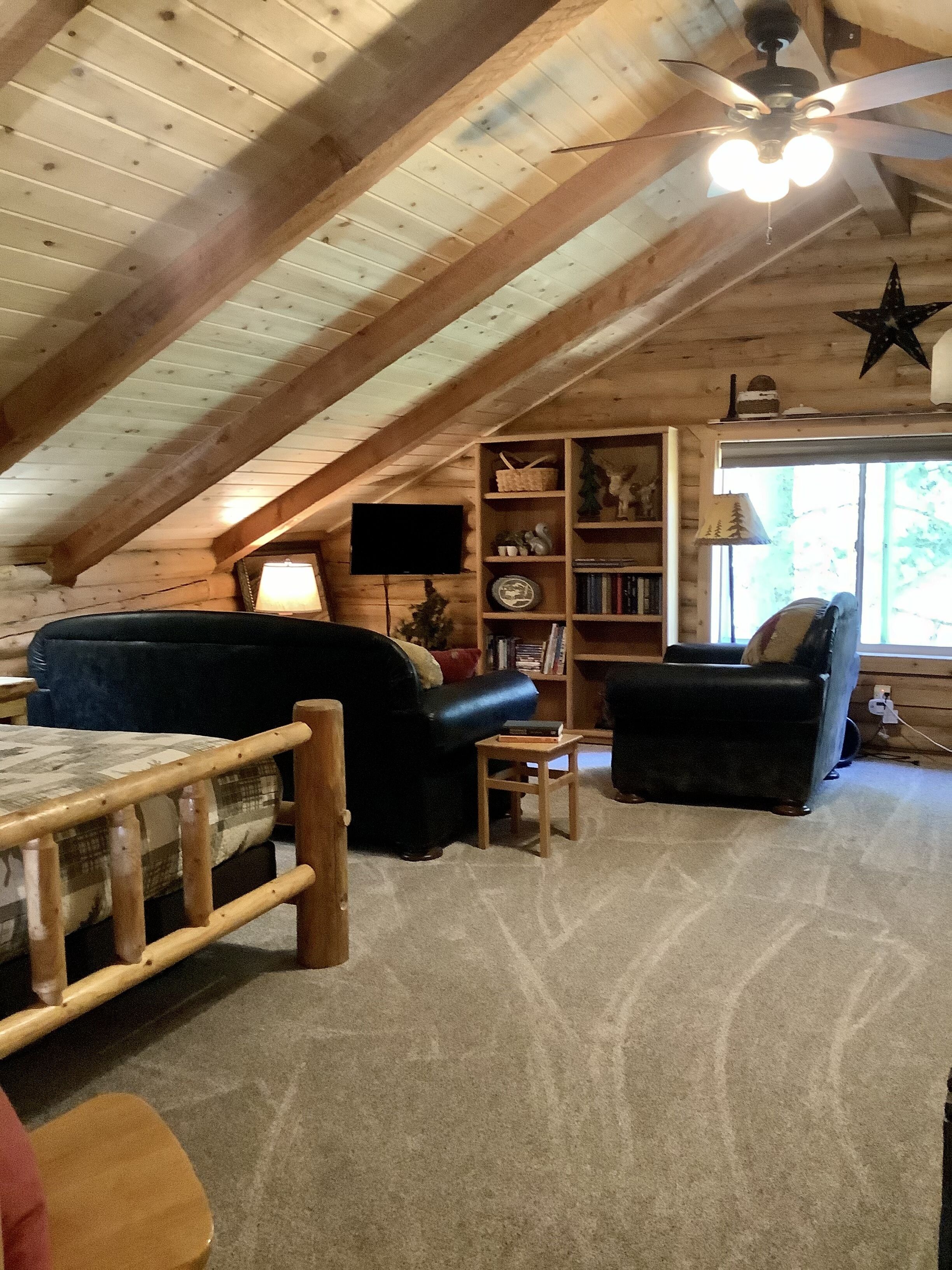 A cozy attic room with a sloped ceiling, wooden beams, a ceiling fan, a star decoration, a bookshelf, a television, and a window with a view of trees.