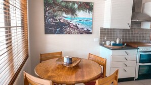 A kitchen with a round wooden table, chairs, and a painting of a beach scene. - Little Beach Bungalow - at Bartletts (Bonny Hills)