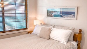 A neatly made bed with striped bedding, a wooden headboard, a bedside lamp, and a framed coastal landscape picture on the wall. - Little Beach Bungalow - at Bartletts (Bonny Hills)
