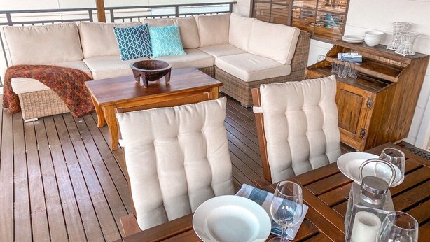 Una terraza de madera con sofá, sillas con cojines y una mesa de comedor dispuesta con platos, vasos y una vela.