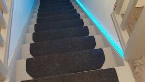 A staircase with black carpeted steps, white walls, and a blue LED light strip along the handrail. - Chambre Avec Douche Privée , Espace Cuisine (Deauville)