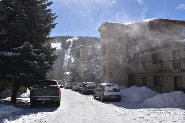 A snowy street with parked cars and buildings, including a multi-story structure with balconies.