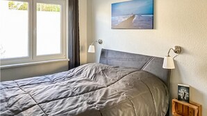 En pænt redt seng med et gråt dynebetræk, et maleri af en strandscene på væggen og en lille træhylde med bøger.