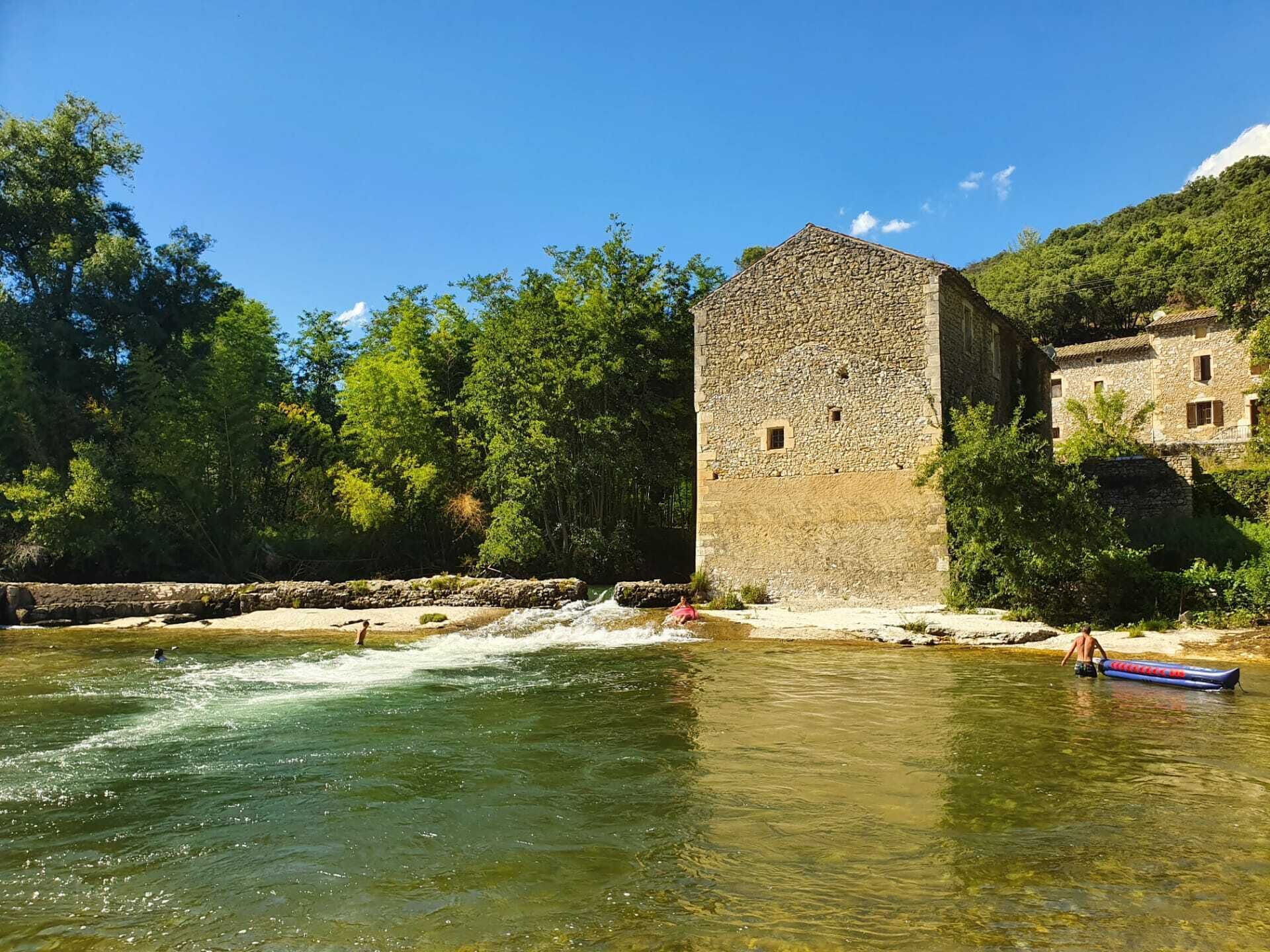 A stone building with a river in front, surrounded by trees and a clear sky.