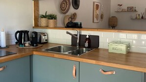 Una cocina moderna con encimera de madera, pileta de acero inoxidable y muebles color esmeralda con detalles en cobre. Se observan lámparas colgantes, platos decorativos y un estante con diversos objetos.