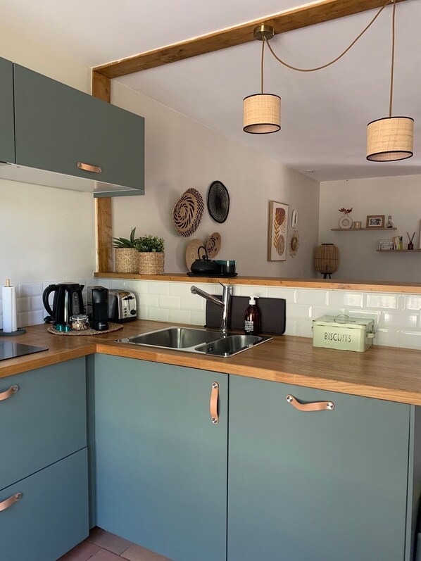Una cocina moderna con encimera de madera, pileta de acero inoxidable y muebles color esmeralda con detalles en cobre. Se observan lámparas colgantes, platos decorativos y un estante con diversos objetos.