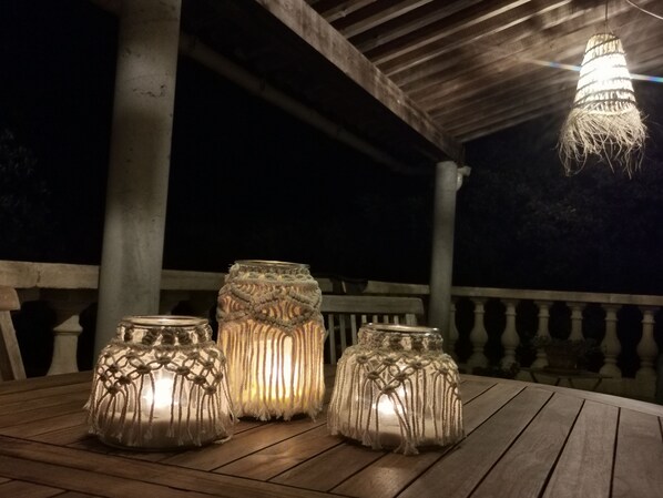 Three macrame candle holders on a wooden table under a wooden pergola at night.