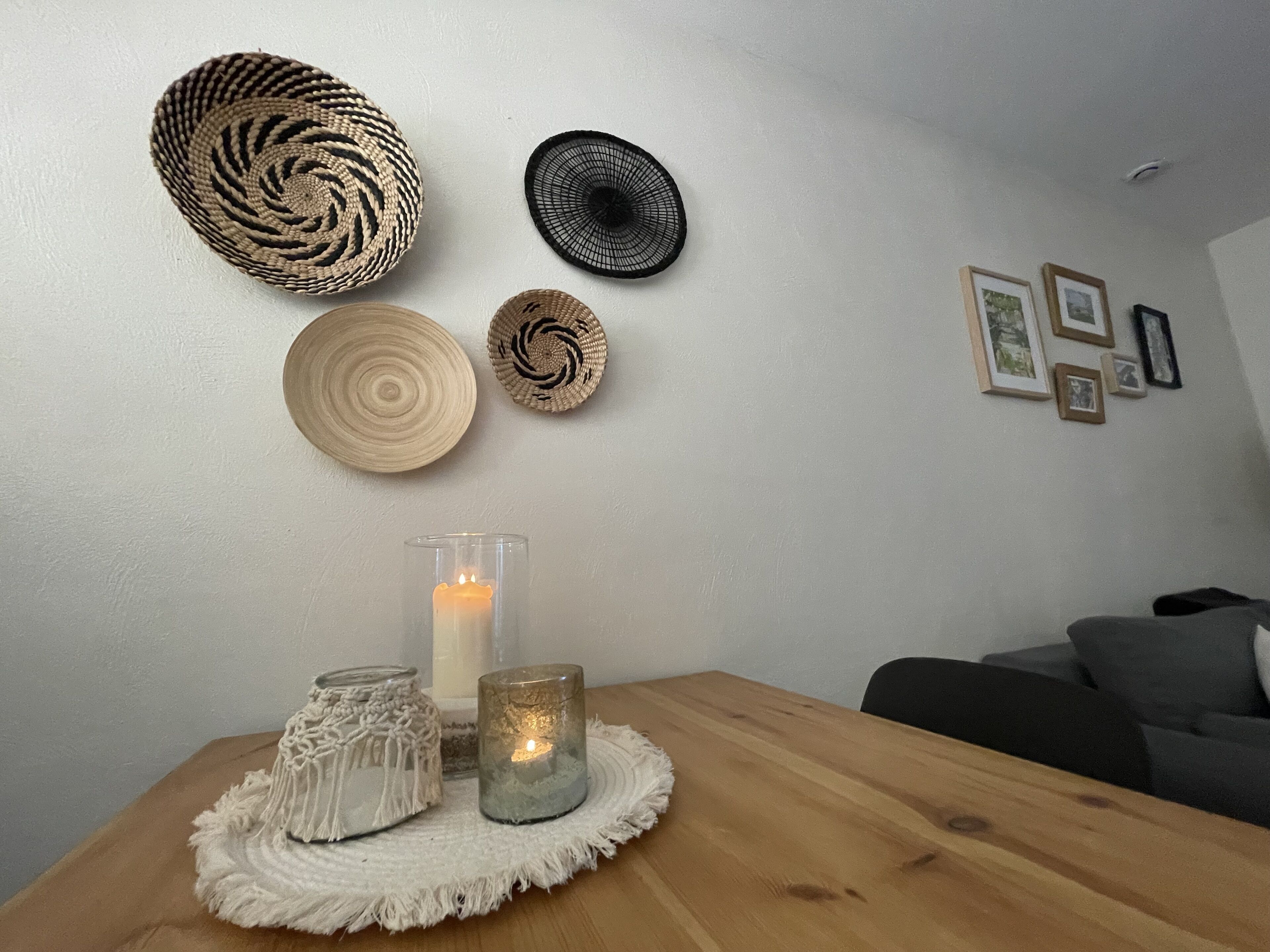 A wooden table with two glass candle holders and a macrame candle holder, a woven wall hanging, and a framed picture collection.