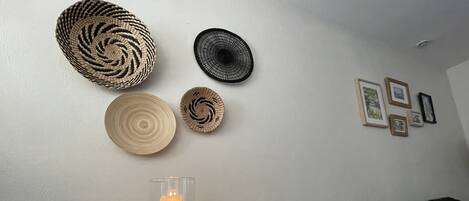 A wooden table with two glass candle holders and a macrame candle holder, a woven wall hanging, and a framed picture collection.