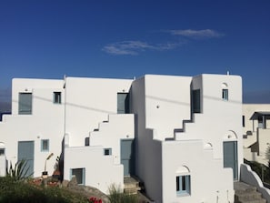A white building with multiple levels and balconies, featuring a clear sky and some greenery in the foreground.