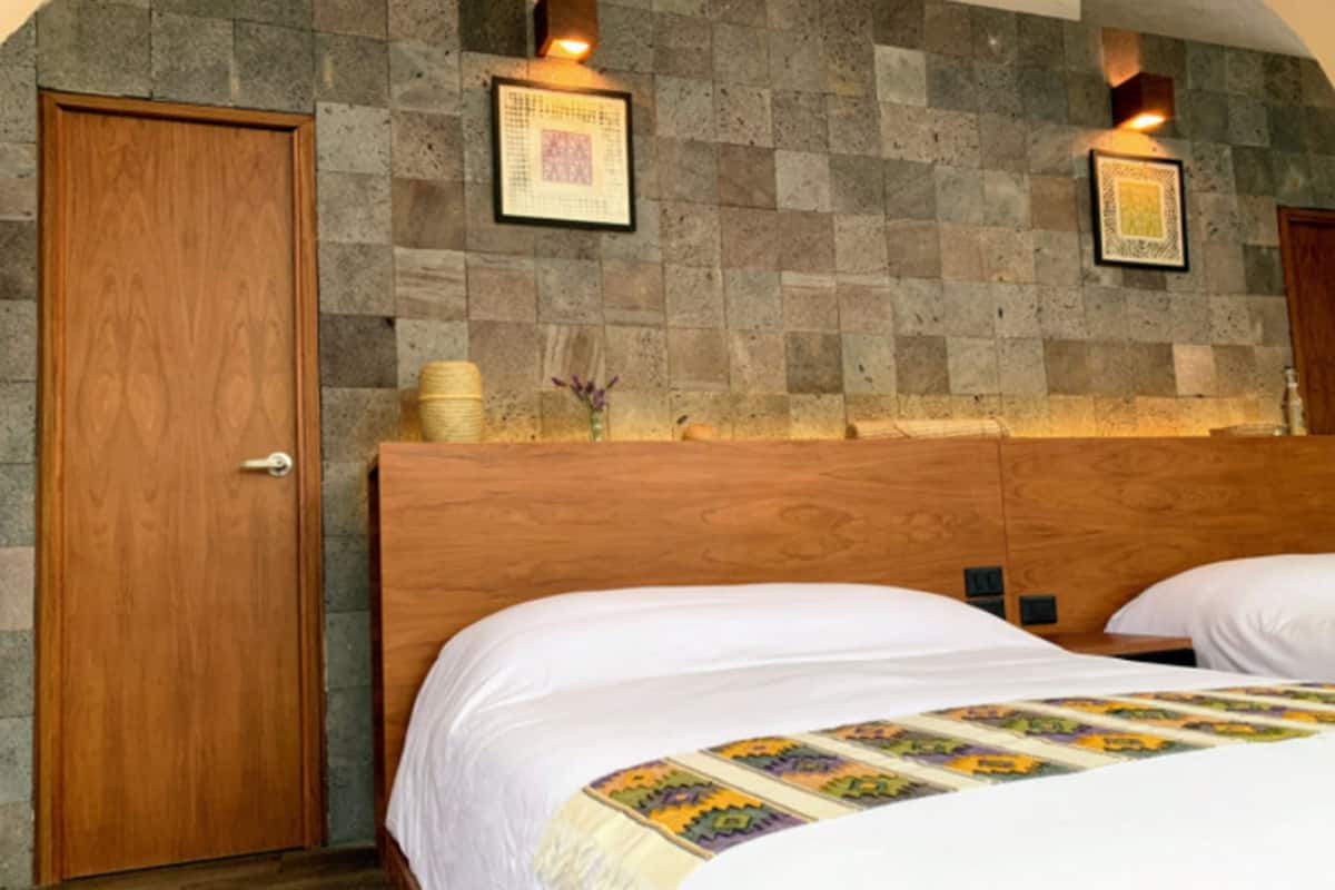 Une chambre d’hôtel avec une porte en bois, deux lits avec du linge de lit blanc, un mur en pierre et deux lampes fixées au mur.