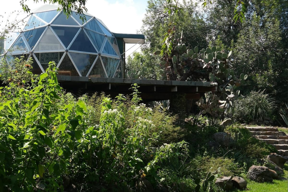 Une structure en forme de dôme géodésique dotée d’une façade en verre, entourée d’une végétation luxuriante et d’un pont en pierre.