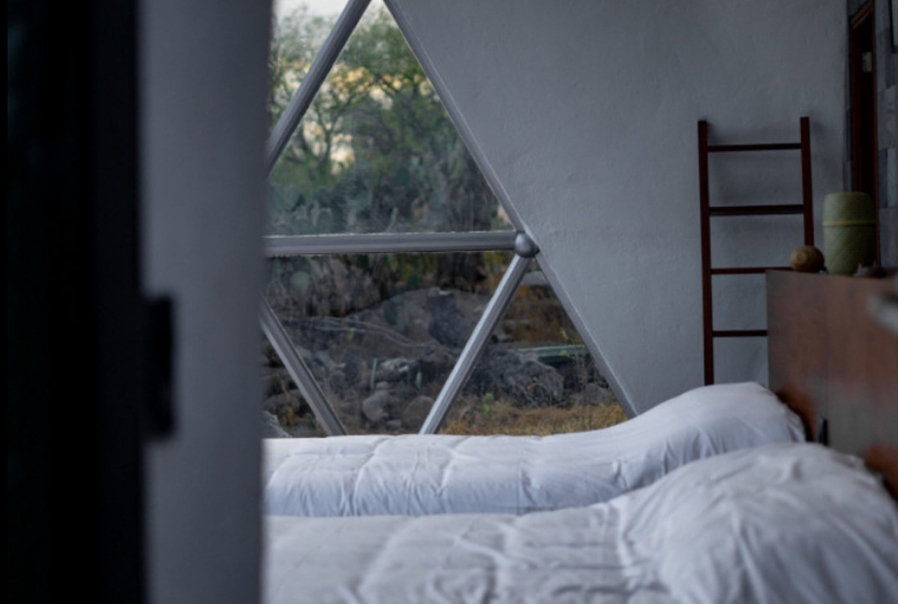 A bed with white bedding, a wooden ladder, and a view of rocks and trees through a triangular window.