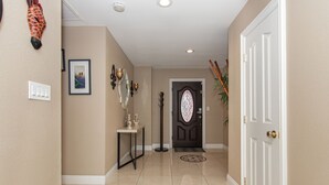 Un pasillo con paredes color beige, una puerta blanca con un panel de vidrio decorativo, una pequeña mesa con una estatua y una lámpara de pie.