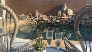 Une chaise en osier, une table en verre, une plante en pot et deux coupes de champagne sont disposés près d’une piscine bordée de rochers, avec un mur en bois en arrière-plan.