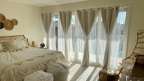 Une chambre à coucher avec un lit, deux poufs, une chaise en osier, un panier en osier et des rideaux en voile.