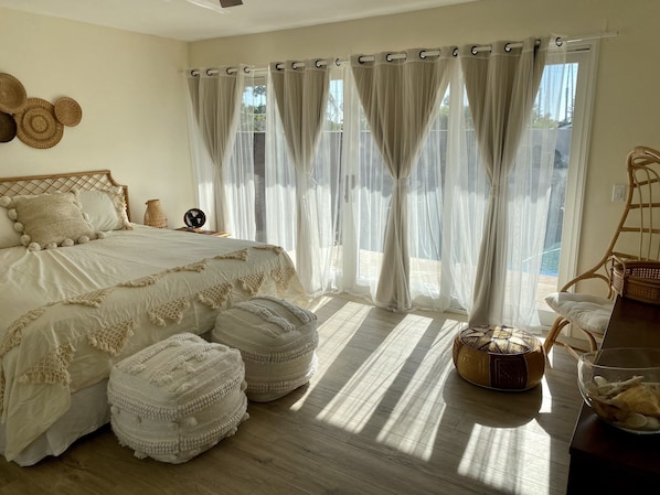 A bedroom with a bed, two ottomans, a wicker chair, and a view of the outdoors.
