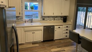 Una cocina con gabinetes blancos, un refrigerador de acero inoxidable, un lavavajillas y un ventanal con vista al patio trasero.
