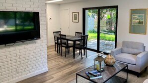 Een moderne woonkamer met een flatscreen televisie bevestigd aan een witte bakstenen muur, een eethoek met een houten tafel en stoelen, een grijze bank en een grote schuifdeur die uitkomt op een achtertuin.