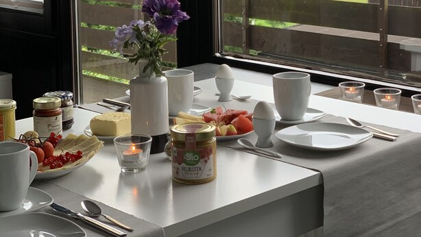 Ein Frühstückstisch ist reichhaltig gedeckt mit Speisen, eine Vase mit Blumen und ein Blick auf eine grüne Landschaft durch große Fenster.