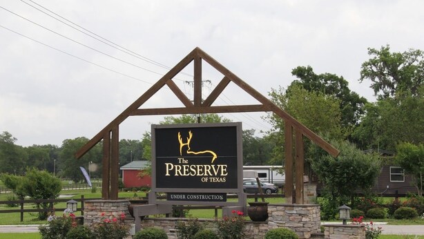 Skilt for "The Preserve of Texas" med et hjortedesign, under konstruktion, omgivet af grøn vegetation og et landligt miljø.