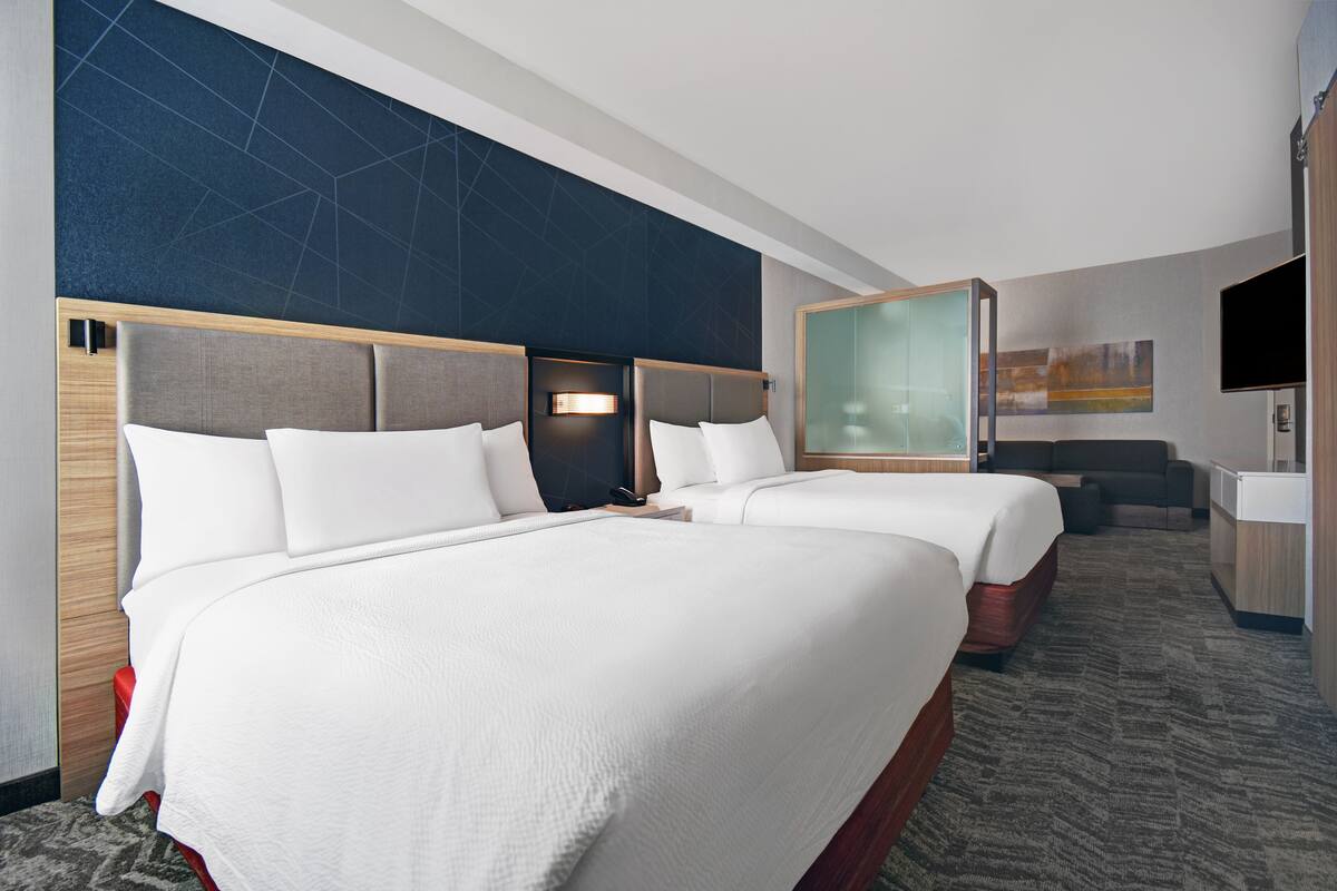 A hotel room with two beds, a desk, a TV, and a glass partition.