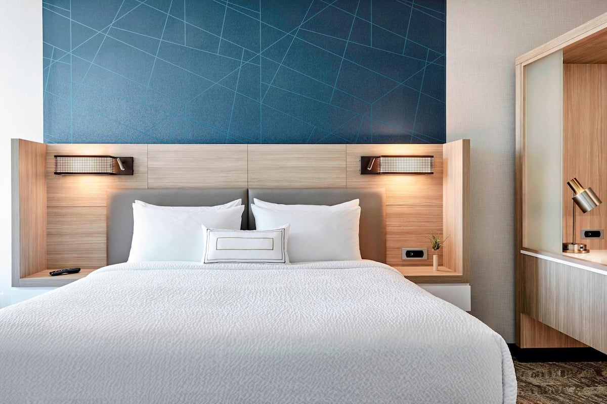 A hotel room with a large bed, two bedside tables, a nightstand, a wooden headboard, and a blue geometric wall design.