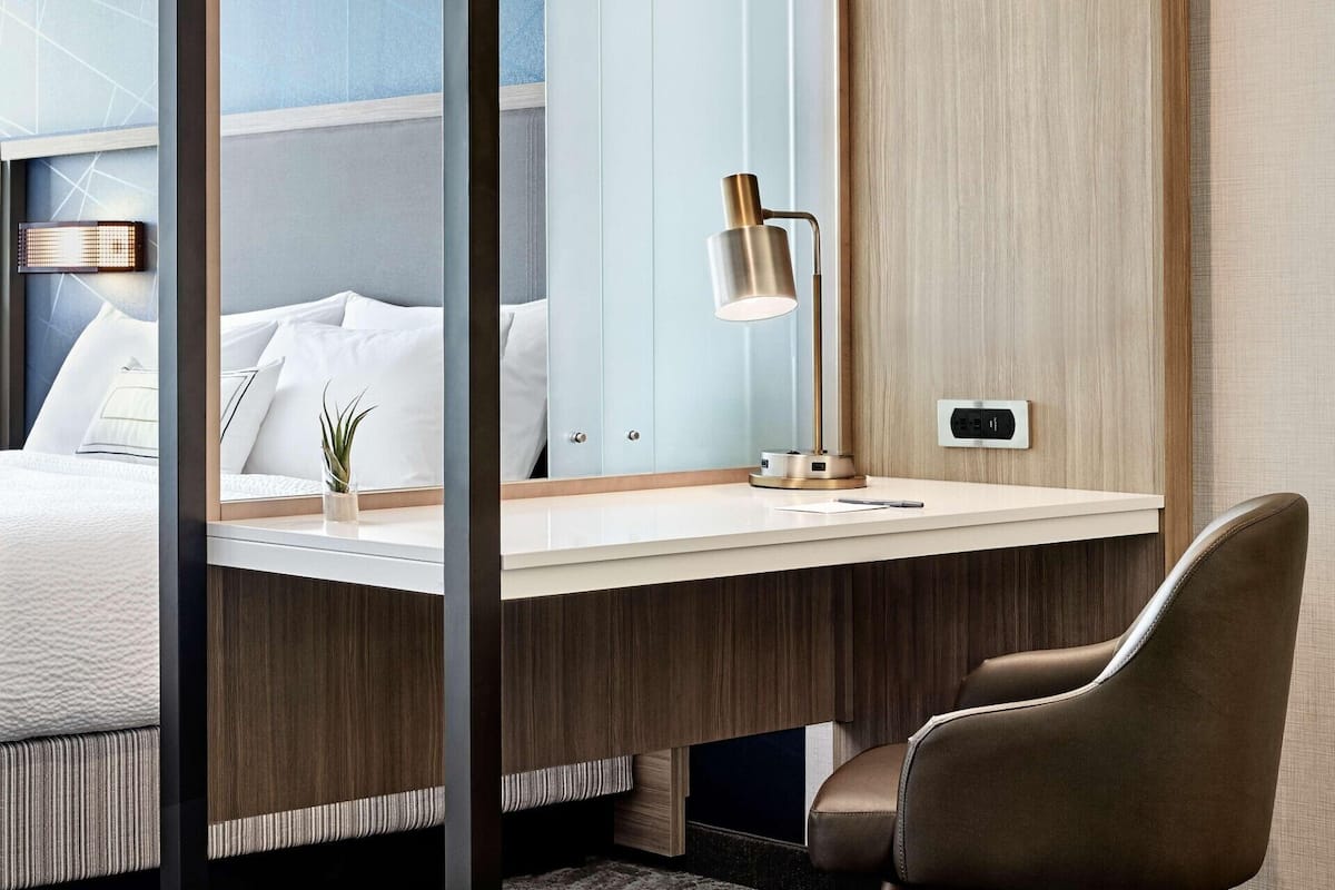 A modern hotel room with a bed, bedside table, lamp, and a chair.