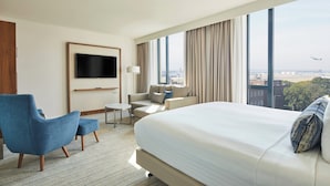 A hotel room with a large bed, a blue armchair, a sofa, a TV mounted on the wall, and a view of the cityscape. - Courtyard by Marriott London Heathrow Airport (Hayes)