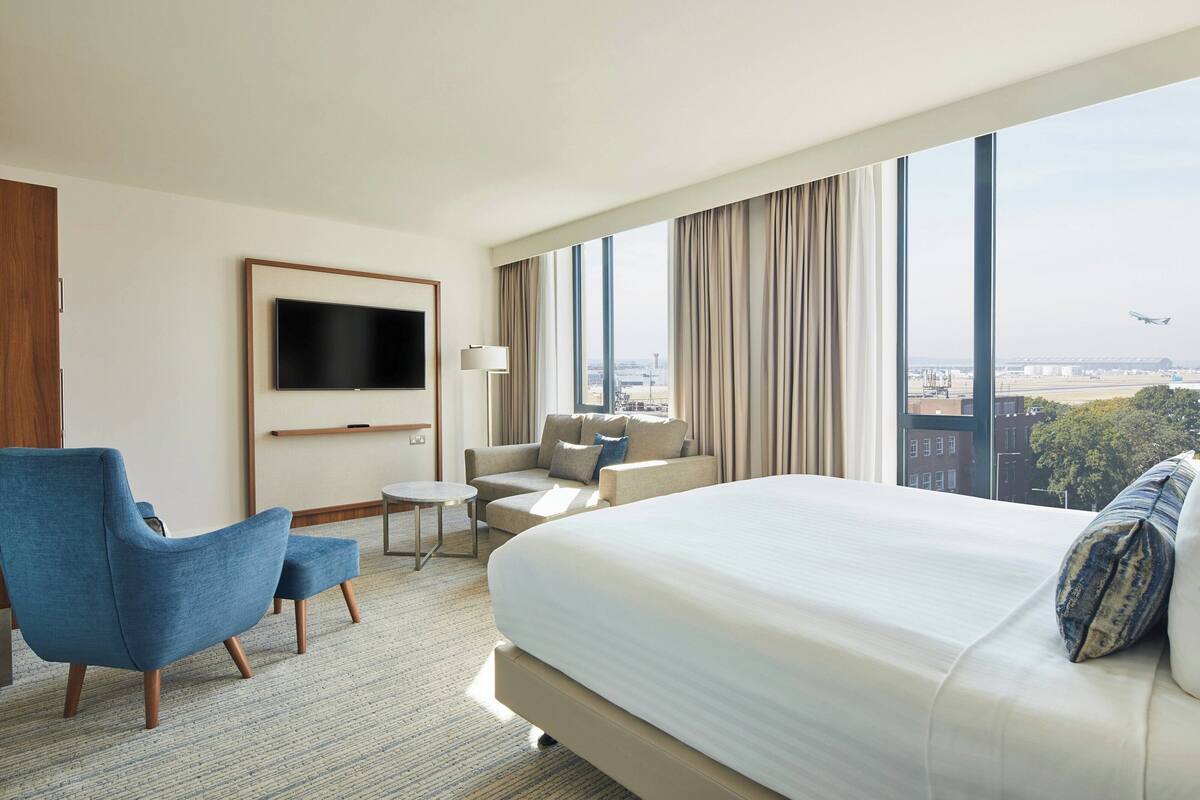 A hotel room with a large bed, a blue armchair, a sofa, a TV mounted on the wall, and a view of the cityscape.
