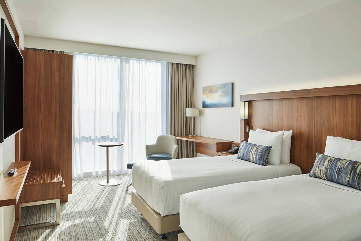A hotel room with two beds, a desk, a chair, a TV, and a window with curtains.