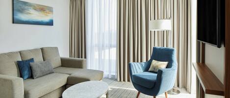Habitación de hotel moderna con un sofá color beige, un sillón azul, una pequeña mesa redonda, un televisor y un amplio ventanal con cortinas.
