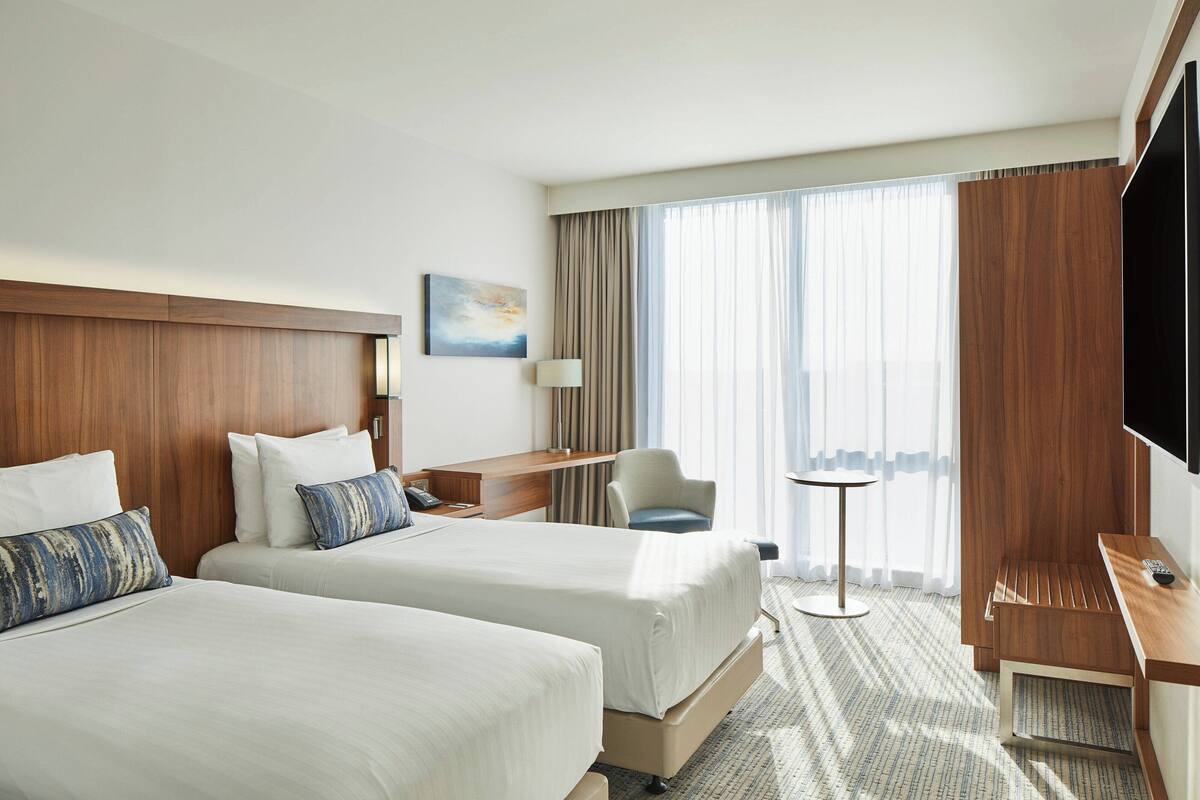 A hotel room with two beds, a desk, a chair, a TV, and a large window with curtains.