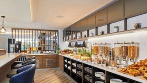 A modern breakfast bar with seating, a variety of food options, and a self-serve beverage station. - Courtyard by Marriott London Heathrow Airport (Hayes)
