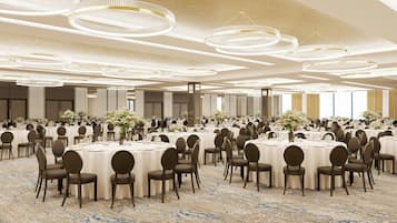 A large banquet hall with round tables and chairs, arranged in rows, with a carpeted floor and multiple chandeliers.