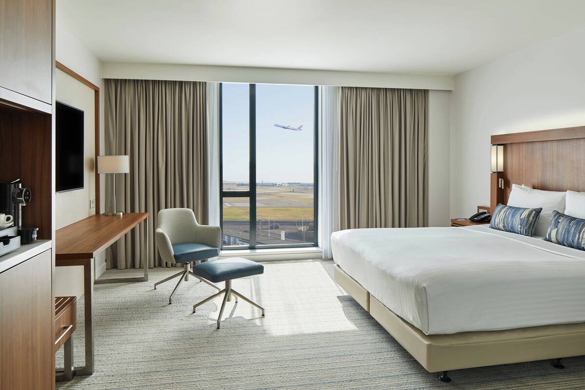 A hotel room with a large bed, a desk, a chair, a TV, and a view of an airplane taking off.